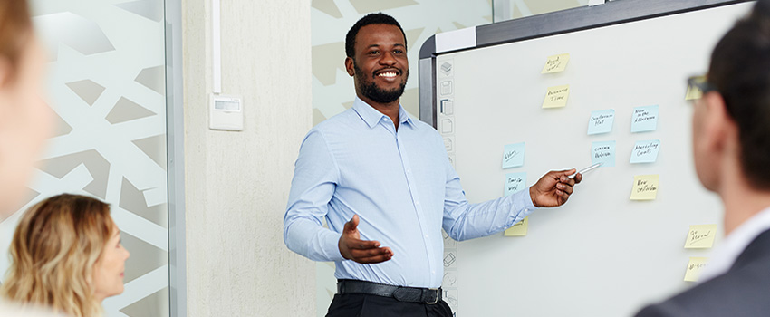 Man speaking in front of white board with people looking on.