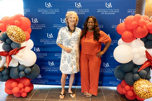 Two people in front of Mitchell College of Business background with balloons.