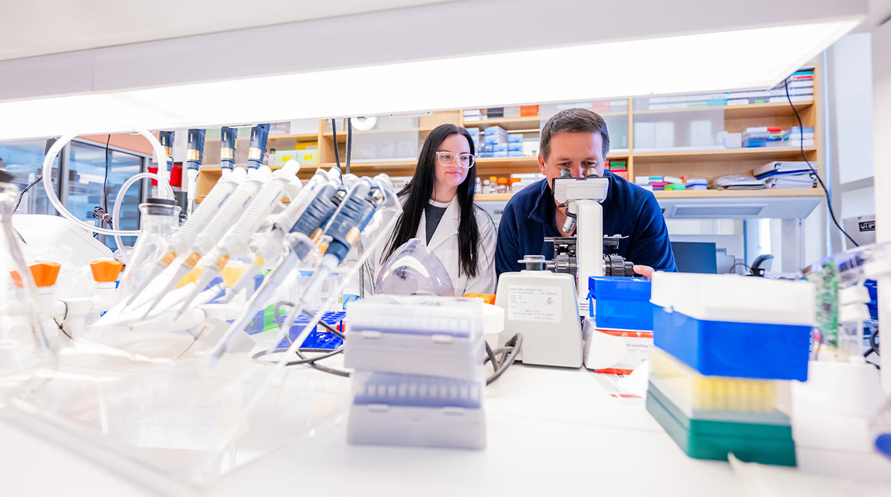 Professor looking in microscope with student looking on in lab.
